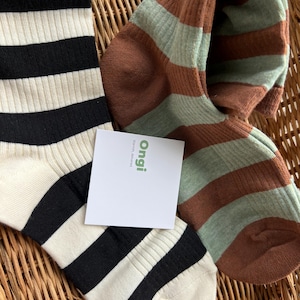 May include: Two pairs of ribbed socks. One pair features horizontal black and cream stripes. The other pair features horizontal brown and sage green stripes. A small white square tag is placed on top, displaying the green text "ongi" and "Warmth, delivered" in smaller black font.