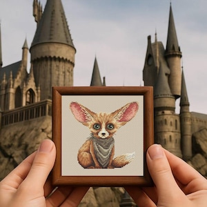 May include: A framed cross-stitch of a fennec fox with large ears, wearing a gray bandana. The fox is brown and white, with green eyes. The frame is a dark wood. The background is a castle.
