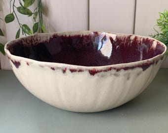 May include: A large, off-white ceramic bowl with a deep burgundy interior and a textured rim. The bowl is set against a neutral background with greenery, suggesting a decorative or serving piece for the home.