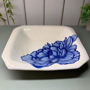 May include: A square, cream-coloured ceramic dish featuring a hand-painted blue floral design. The dish has a slightly raised edge and a smooth surface. The blue flower is detailed and covers a portion of the dish.