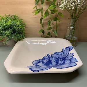 May include: A beige, square ceramic serving dish with a hand-painted blue floral design. The dish has a slightly raised rim and is suitable for serving food or as a decorative piece. The floral design is a prominent feature.