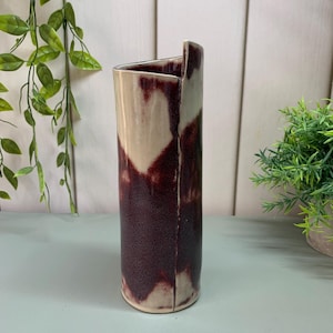 May include: A tall, ceramic vase with an unusual, angular design. The vase has a speckled, deep burgundy colour that fades to a creamy beige. The vase is approximately 25 cm tall, ideal for displaying flowers or as a decorative piece.