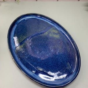 May include: Oval ceramic serving platter with a glossy, deep blue glaze. The handmade dish has a slightly raised rim and a speckled, textured surface. The colour transitions from a lighter blue at the edges to a darker, almost indigo, centre.