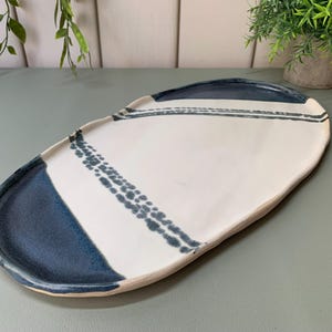 May include: An oval ceramic serving platter with a smooth off-white centre and deep blue glazed edges. The platter features two distinct dark blue decorative bands across the surface, one solid and one composed of small, textured dotted patterns.