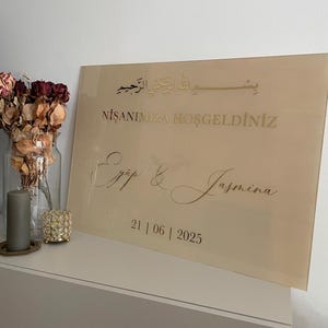 May include: A rectangular, beige acrylic sign with gold lettering. The sign includes Arabic script, the words "NİŞANIMIZA HOŞGELDİNİZ", and the names "Ey&uuml;p & Jasmina" along with the date "21 | 06 | 2025".