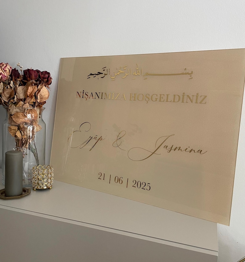 May include: A rectangular, beige acrylic sign with gold lettering. The sign includes Arabic script, the words "NİŞANIMIZA HOŞGELDİNİZ", and the names "Ey&uuml;p & Jasmina" along with the date "21 | 06 | 2025".