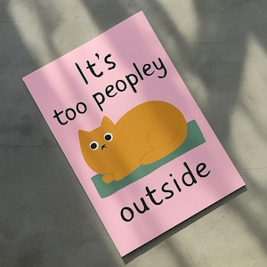 Könnte beinhalten: Eine rosa Karte mit dem Text "It's too peopley outside" in Schwarz. Eine orangefarbene Katzenillustration befindet sich auf einer grünen Matte. Die Karte ist abgewinkelt, mit Schatten auf der Oberfläche.