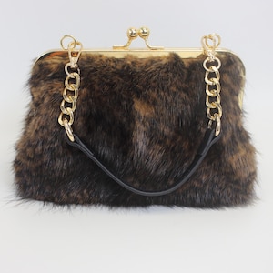 May include: A brown and black faux fur handbag with a gold-tone clasp and chain strap. The bag has a rectangular shape and a black leather handle. The clasp is a kiss-lock style.