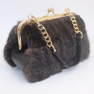 Op de afbeelding: Een bruine bont handtas met een goudkleurige sluiting en kettingriem. De tas heeft een rechthoekige vorm met een afgeronde bovenkant en een zwarte leren basis. De kettingriem is bevestigd aan de tas met goudkleurige hardware.