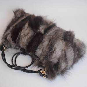 May include: A silver and black fur handbag with a black leather strap and gold-coloured hardware. The bag has a soft, textured appearance and a rounded shape. The strap is looped and attached with small gold clasps.