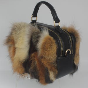 May include: A black leather handbag with a top handle and gold-colored hardware. The bag features a fluffy fur trim in shades of brown, tan, and cream. The bag has two zippered compartments.