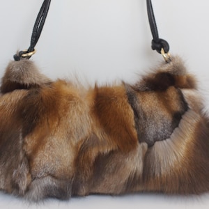 Peut inclure: Un sac à main rectangulaire en fourrure marron, beige et grise. Le sac est doté d'une bandoulière en cuir noir avec des anneaux en métal doré. La fourrure semble être du renard ou d'un animal similaire. Le sac est de couleur neutre.