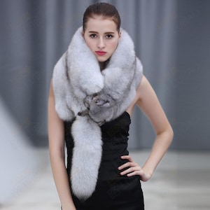Puede incluir: Una lujosa bufanda de piel de color gris claro con una larga cola fluida. La bufanda está drapeada alrededor del cuello y presenta un diseño detallado y texturizado en la parte delantera. La piel parece suave y esponjosa, creando una pieza llamativa.