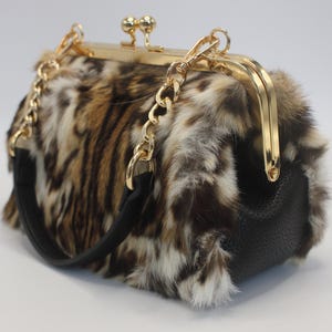 May include: A rectangular handbag with a faux fur exterior in shades of brown, tan, and white. The bag features a gold-tone clasp and chain strap, with a black leather handle and base. The bag is approximately 8 inches long.
