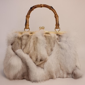 Puede incluir: Un bolso de mano vintage con asa de bambú y cierre dorado. El bolso está cubierto con una mezcla de pieles blancas y marrón claro, creando una apariencia texturizada. El asa se arquea elegantemente en la parte superior.