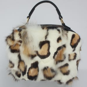 May include: A handbag with a black handle and a zipper closure. The bag is covered in a white, brown, and black leopard print fur. The handle is black and attached with gold-colored hardware.