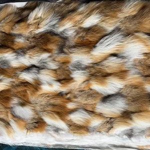May include: A rectangular piece of fox fur in shades of brown, white, and gray. The fur has a dense, textured appearance, with the individual hairs creating a soft, layered effect. The fur is likely used for clothing or decorative purposes.