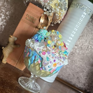 May include: A decorative glass with a colorful, ornate design, filled with an assortment of beads, gems, and pearls. A vintage book, a small bear figurine, and a bouquet of flowers are also present. A gold spoon rests on top of the glass.