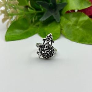 Könnte beinhalten: Ein silberfarbener Metallring mit einem detaillierten Eidechsen- oder Gecko-Motiv auf der Oberseite. Das Eidechsendesign hat eine texturierte, oxidierte Oberfläche mit kleinen schwarzen Akzenten entlang des Körpers und des Schwanzes.