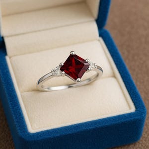 May include: A silver ring with a square, deep red gemstone as the centerpiece. The ring is flanked by smaller, clear stones on each side. The ring is displayed in a blue velvet jewelry box.