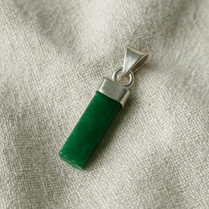 Könnte beinhalten: Rechteckiger Anhänger mit einem leuchtend grünen Stein, akzentuiert durch eine silberfarbene Metallkappe und Öse. Der Anhänger wird auf einem neutralfarbenen Stoff präsentiert, der seine polierte Oberfläche und sein schlichtes Design hervorhebt.