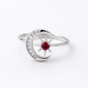 May include: A silver ring featuring a crescent moon design encrusted with baguette-cut stones. A star with a red gemstone sits in the center. The ring has a polished finish and is set against a white background.