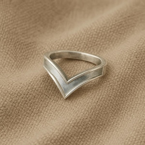 Könnte beinhalten: Ein silberner Ring mit V-förmigem Design. Der Ring hat ein flaches Band und eine strukturierte Oberfläche, die auf einem beigefarbenen Stoffhintergrund liegt. Das Design des Rings ist schlicht und modern.