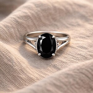 Könnte beinhalten: Ein silberner Ring mit einem großen, oval geschliffenen schwarzen Edelstein. Der Ring hat ein geteiltes Schaftdesign und liegt auf einem neutralfarbenen Stoff. Der polierte Edelstein ist der Blickfang.