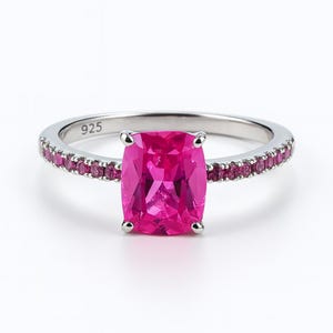 May include: A silver ring with a large, square, hot pink gemstone as the focal point. The band is adorned with smaller pink gemstones. The ring is set against a white background, with the number "925" visible on the band.