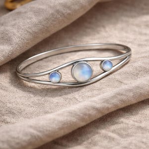 Peut inclure: Un bracelet argenté orné de trois cabochons ronds en pierre de lune irisée. Le bracelet présente un design délicat et ouvert avec les pierres disposées en ligne. Les pierres ont un aspect doux et laiteux. Le bracelet est présenté sur un tissu de couleur neutre.