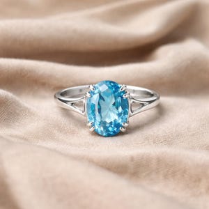 May include: A silver ring with a large, oval-shaped, light blue gemstone. The gemstone is the central feature, held in place by prongs. The ring's band has a simple, elegant design, with a split shank. The background is a soft, neutral fabric.