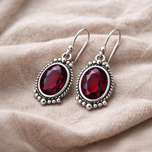 May include: A pair of silver-toned drop earrings with oval, deep red gemstones. The gemstones are set in a decorative frame with a beaded border. The earrings are displayed on a beige fabric background.