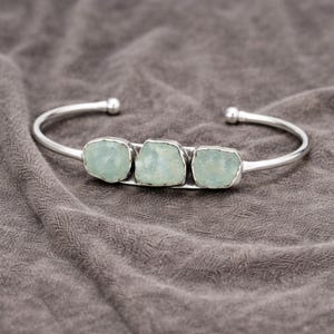 May include: A silver cuff bracelet with three pale blue aquamarine stones. The bracelet has a simple, open design with rounded ends. The stones are set in individual bezels. The bracelet's design is minimalist and elegant.