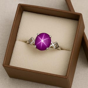 May include: A sterling silver ring featuring a large, oval-cut, purplish-pink star sapphire gemstone. The ring has a classic design with a six-pointed star effect within the stone and geometric accents on the band. The ring is presented in a brown jewelry box.