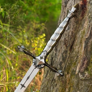 May include: A silver-toned metal sword featuring a spiralled handle, a pointed pommel, and a crossguard with angular, geometric detailing. The blade is straight with a central fuller and a distinct cross-shaped engraving near the hilt.