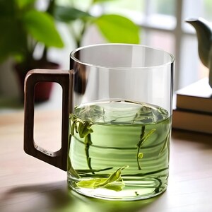 Pode incluir: Uma caneca de vidro transparente com uma al&ccedil;a de madeira escura, cheia de ch&aacute; verde e folhas de ch&aacute;. A caneca est&aacute; em uma superf&iacute;cie de madeira, com um fundo desfocado de plantas e uma chaleira.