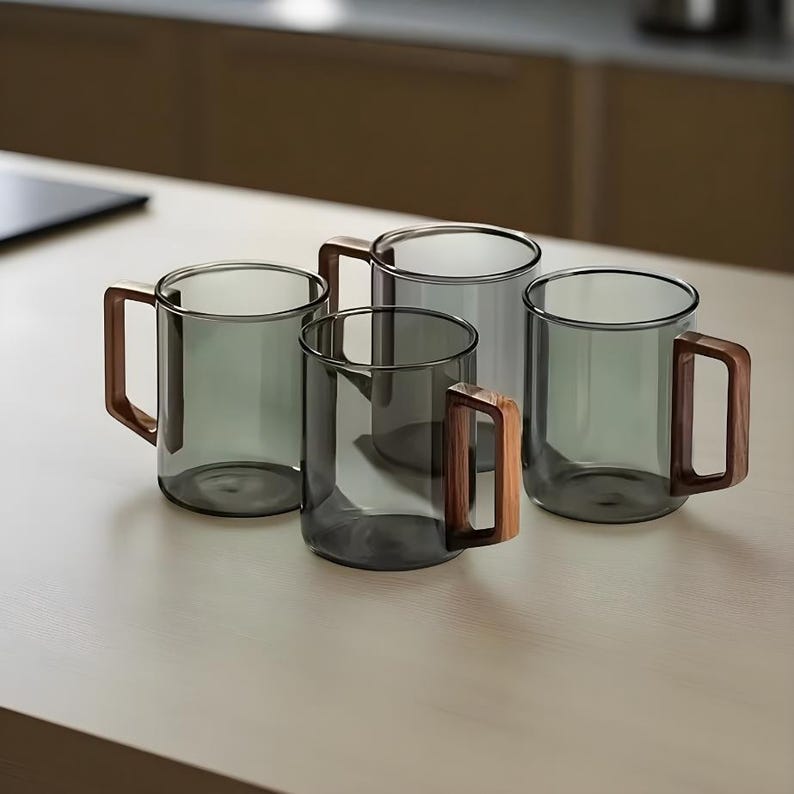 Pode incluir: Quatro canecas de vidro transparente com um tom cinza esfuma&ccedil;ado e al&ccedil;as de madeira dispostas sobre uma superf&iacute;cie clara. As canecas t&ecirc;m um design simples e moderno, adequado para caf&eacute; ou ch&aacute;. As al&ccedil;as de madeira proporcionam um contraste quente.