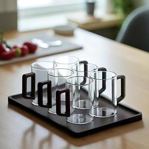 Pode incluir: Um conjunto de seis canecas de vidro transparente com al&ccedil;as castanhas escuras, apresentadas numa bandeja retangular a condizer. As canecas s&atilde;o cil&iacute;ndricas e parecem vazias. A bandeja e as canecas est&atilde;o sobre uma superf&iacute;cie de madeira clara.