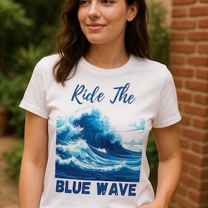 May include: White t-shirt featuring a blue wave design with the text "Ride The Blue Wave". The graphic depicts a stylized ocean wave. The shirt is worn with blue jeans.