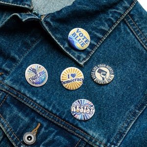 May include: Five circular pin-back buttons attached to a dark blue denim jacket. The buttons feature various designs and text including VOTE BLUE, FIGHT THE OLIGARCHY, I LOVE DEMOCRACY, VOTE LIKE YOUR RIGHTS DEPEND ON IT, and RESIST in blue, white, and gold.