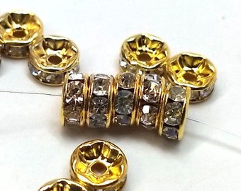 May include: A group of gold-toned metal beads with clear rhinestones. The beads are round and have a raised design. Some of the beads are stacked together to form a longer bead.