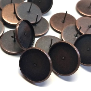 May include: A group of 15 copper-toned earring settings with black backing and a post for attaching earrings. The settings are round and have a smooth surface.