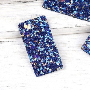 May include: Three rectangular blue glitter shapes with iridescent glitter. The shapes are on a white wooden surface.