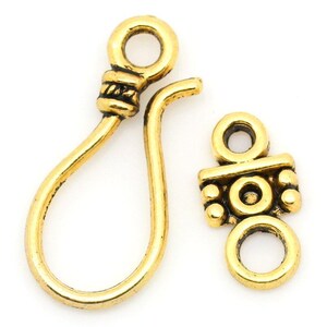 May include: Two antique gold-colored jewelry clasps. One is a hook-and-eye style, with a loop and a curved hook. The other is a decorative rectangular piece with a loop and a central circle, and smaller circles on each side.
