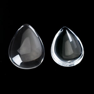 May include: Two clear glass teardrop-shaped cabochons, one larger and one smaller, on a black background.