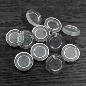 May include: A group of ten clear plastic earring posts with a round base. The posts are arranged in a random pattern on a dark wood surface.