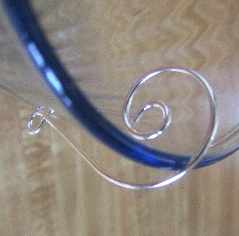K&ouml;nnte beinhalten: Eine silberne Haarklammer aus Draht mit einem Spiraldesign. Die Klammer wird vor einem blau-wei&szlig;en Hintergrund gezeigt.