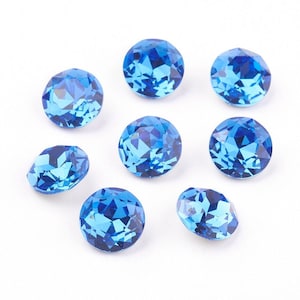 Puede incluir: Ocho gemas facetadas, redondas y ovaladas, de color azul brillante, dispuestas sobre una superficie blanca. Las gemas tienen múltiples facetas que reflejan la luz, creando un efecto brillante. Las gemas probablemente se utilizan para joyería o proyectos de artesanía.