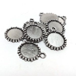 Op de afbeelding: Een verzameling zilverkleurige, ronde sieraden bezels met een getextureerd oppervlak. Elke bezel heeft een kralenrand en een kleine lus om aan een ketting of een ander sieradenonderdeel te bevestigen. De bezels zijn verspreid op een wit oppervlak.