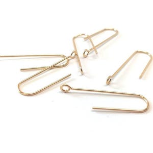 May include: Gold-toned earring hooks, a type of jewelry finding, are shown scattered on a white surface. The hooks are made of metal and have a loop at one end and a hook at the other.